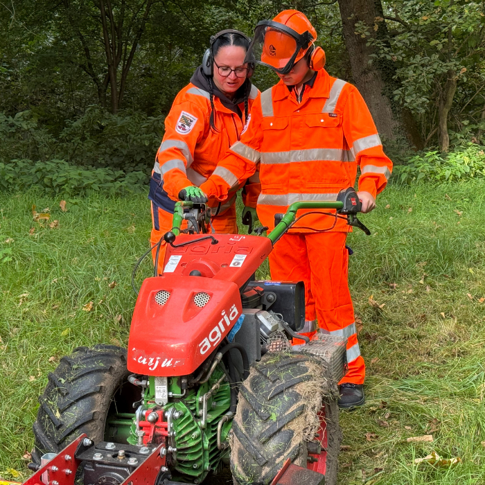 Praktikant und Straßenwärterin stehen hinter einem Rasenmäher. 